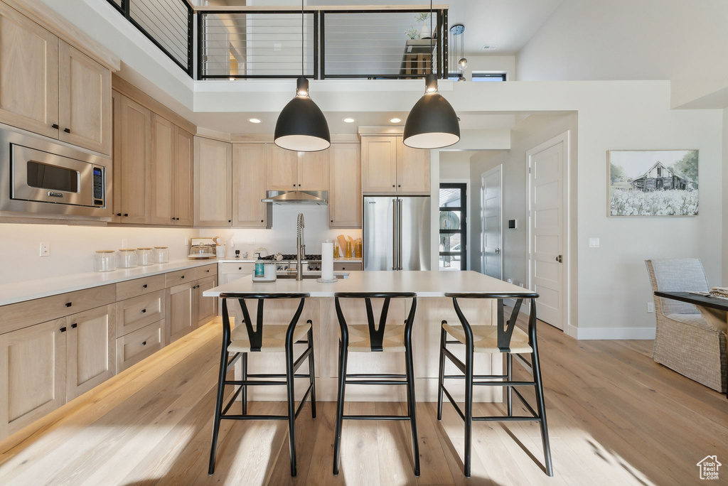 Kitchen with appliances with stainless steel finishes, light wood-type flooring, a towering ceiling, light brown cabinetry, and decorative light fixtures