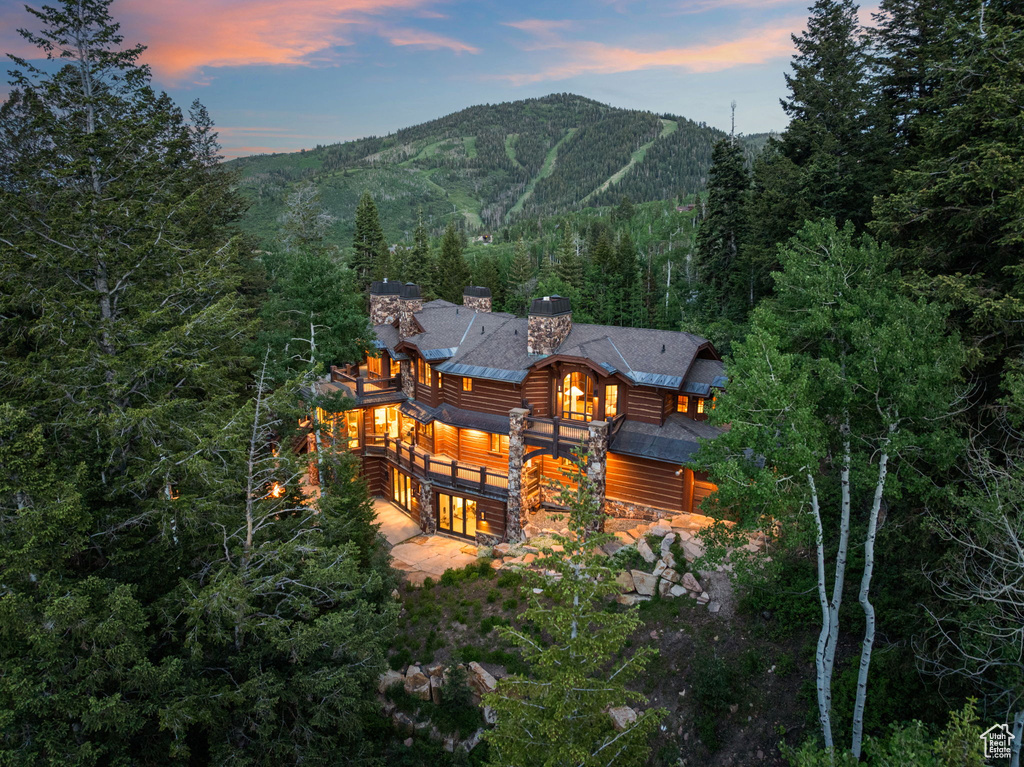 Back of house at dusk featuring a view of trees, a mountain view, stone siding, a balcony, and log siding
