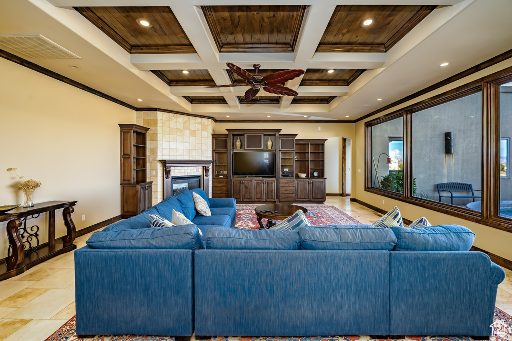 Living room featuring coffered ceiling, crown molding, a tiled fireplace, ceiling fan, and beamed ceiling