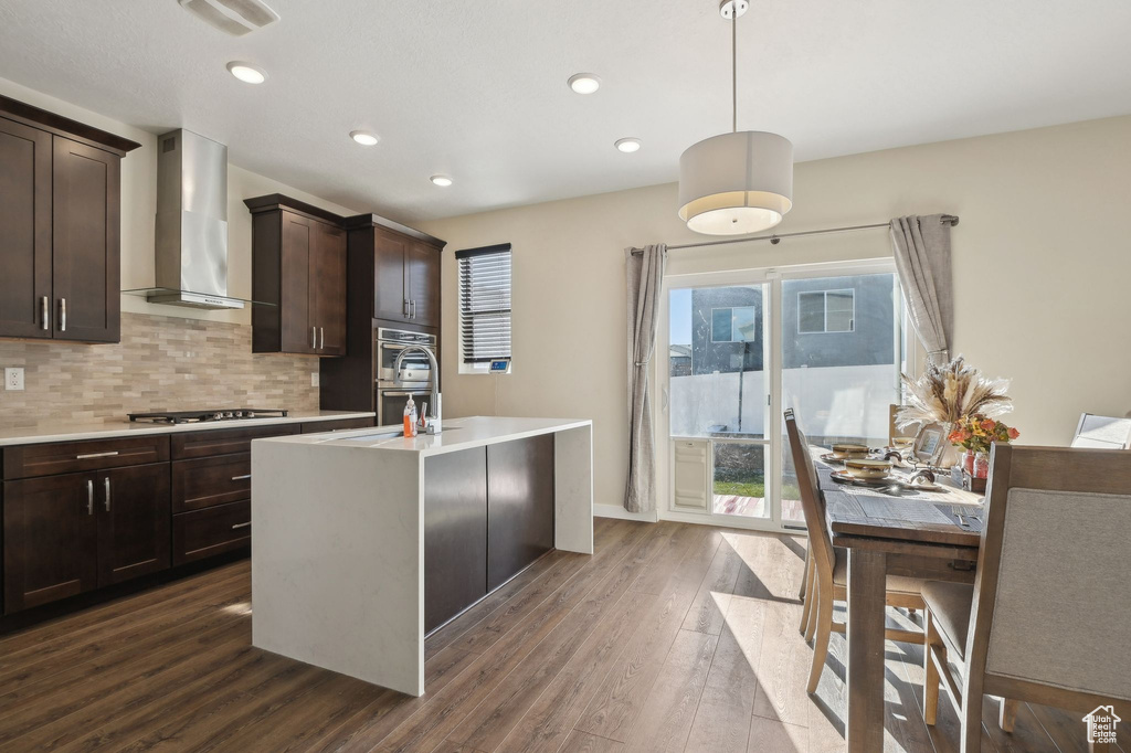 Kitchen featuring stainless steel gas stovetop, wall chimney range hood, pendant lighting, a center island with sink, and dark wood-type flooring