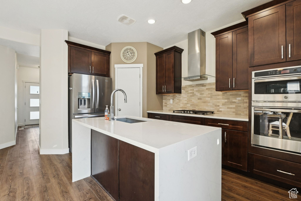 Kitchen with appliances with stainless steel finishes, dark hardwood / wood-style floors, tasteful backsplash, a kitchen island with sink, and wall chimney exhaust hood