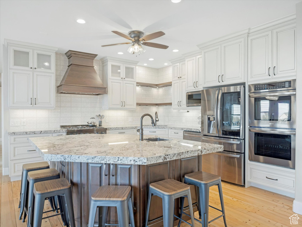 Kitchen featuring a kitchen island with sink, appliances with stainless steel finishes, premium range hood, white cabinets, and sink