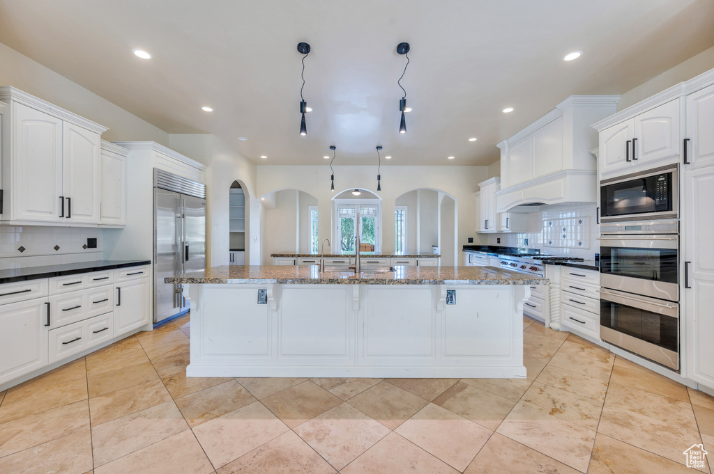 Kitchen with decorative backsplash, built in appliances, a kitchen island with sink, and dark stone countertops