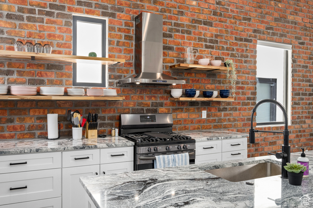 Kitchen with brick wall, wall chimney exhaust hood, sink, light stone counters, and stainless steel range with gas stovetop