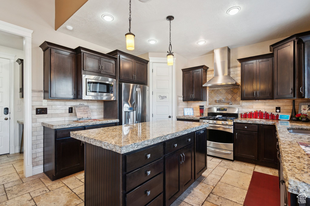 Kitchen featuring wall chimney range hood, appliances with stainless steel finishes, hanging light fixtures, dark brown cabinetry, and a kitchen island