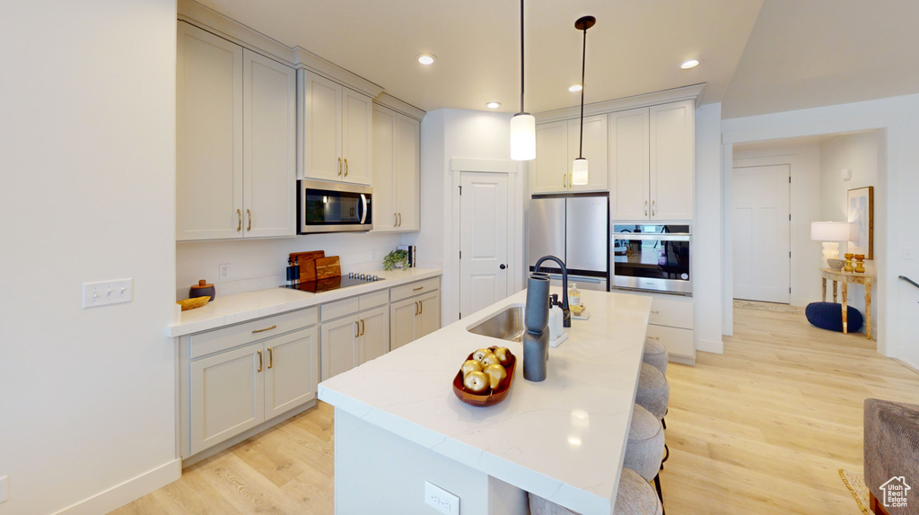 Kitchen with stainless steel appliances, light wood-style flooring, a kitchen island with sink, a sink, and hanging light fixtures