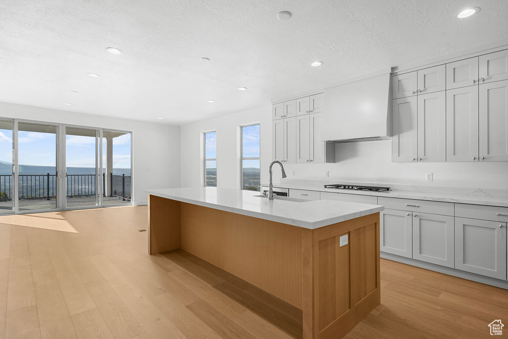 Kitchen featuring wall chimney range hood, sink, light hardwood / wood-style flooring, an island with sink, and white cabinets