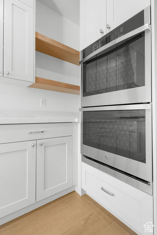 Kitchen with a textured ceiling, stainless steel double oven, white cabinetry, light wood-type flooring, and light stone counters