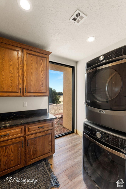 Clothes washing area with stacked washer and dryer, a textured ceiling, cabinets, and light hardwood / wood-style flooring