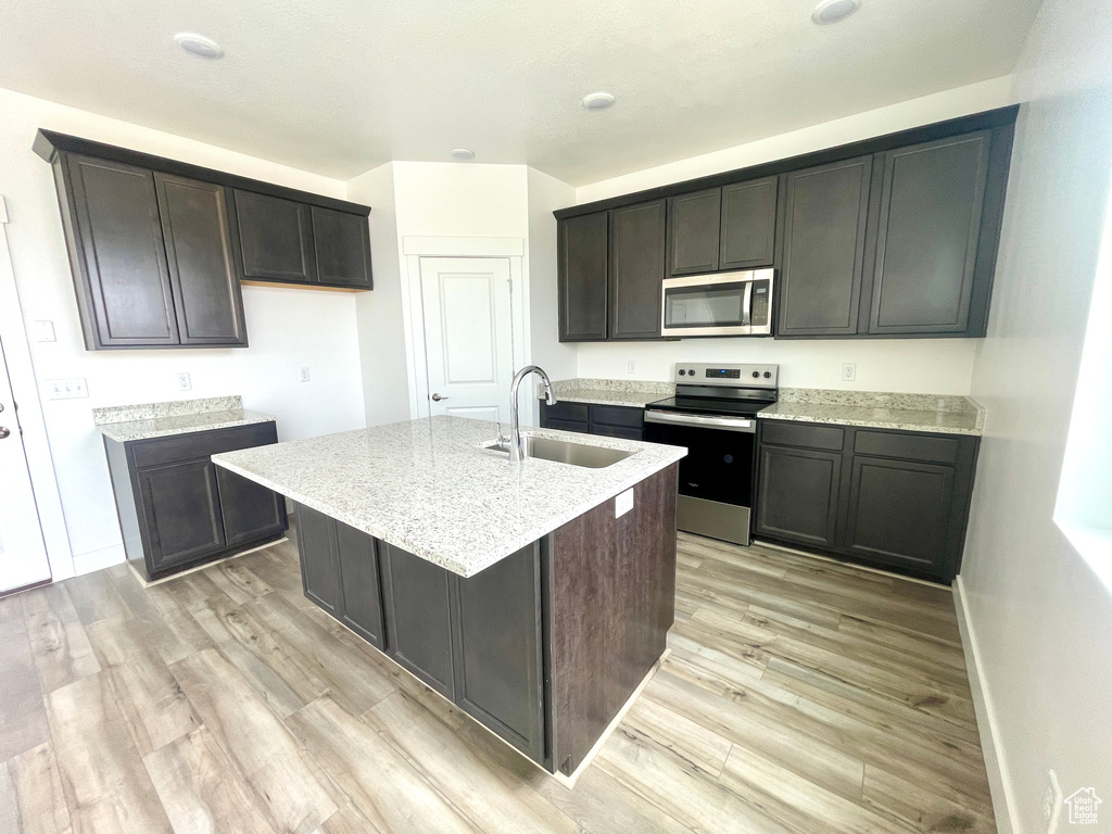 Kitchen featuring light wood finished floors, a kitchen island with sink, stainless steel appliances, a sink, and light stone counters