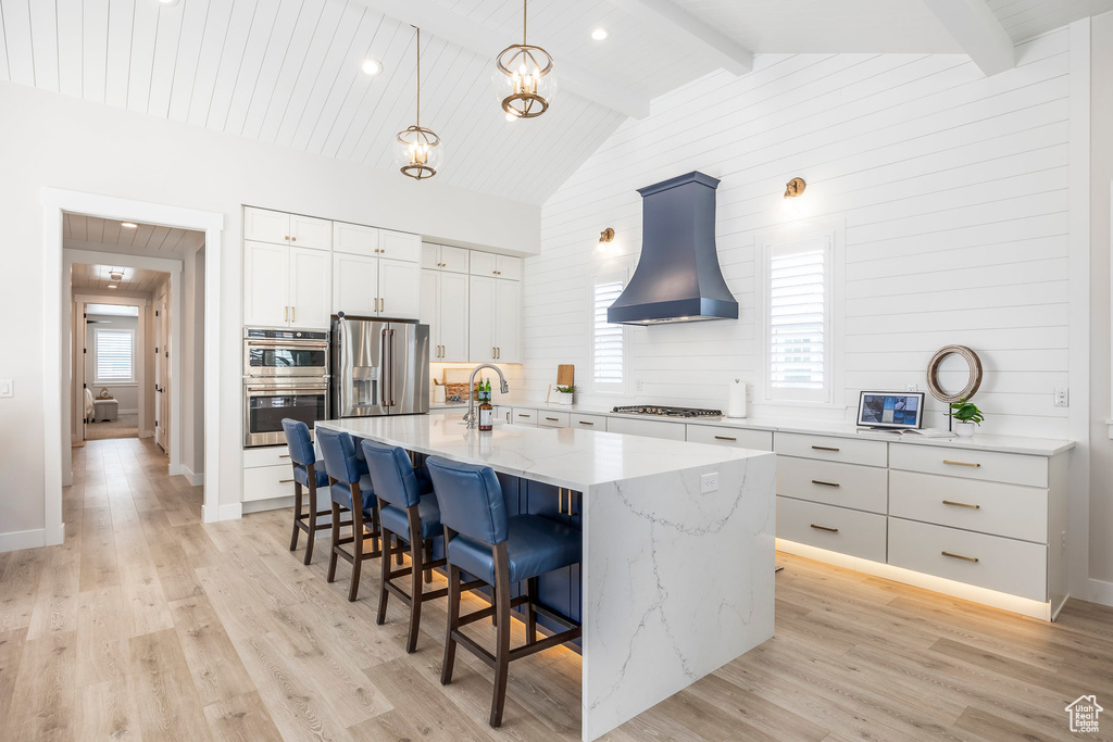 Kitchen with premium range hood, appliances with stainless steel finishes, decorative light fixtures, white cabinetry, and an island with sink