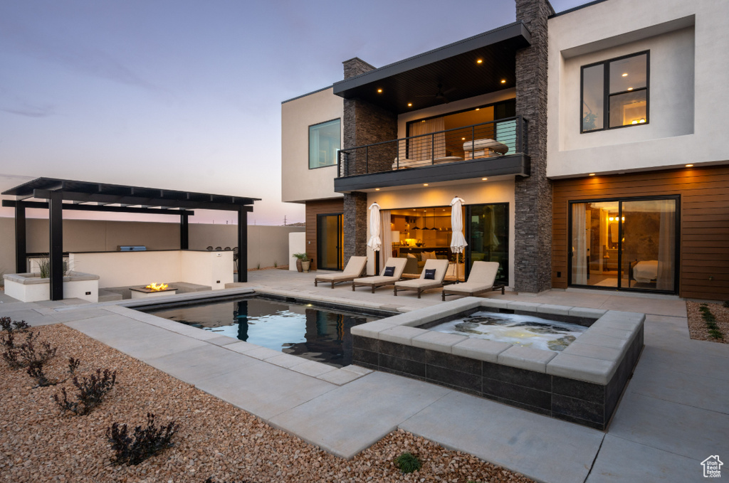 Back of house at dusk with a balcony, an in-ground hot tub, a patio, stone siding, and an outdoor pool