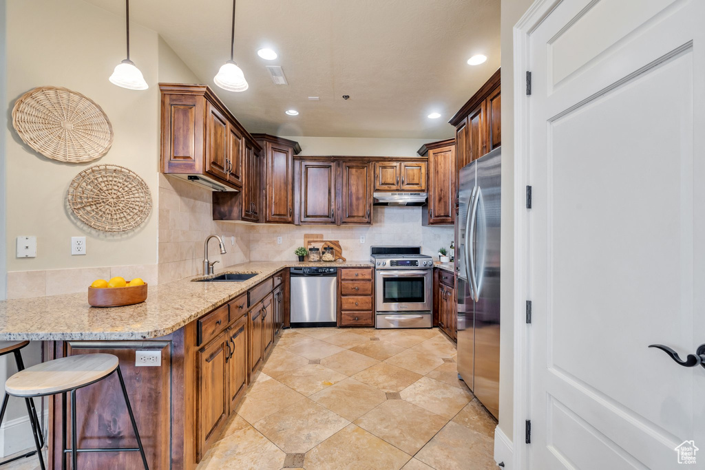 Kitchen with sink, a breakfast bar area, appliances with stainless steel finishes, light stone counters, and decorative light fixtures