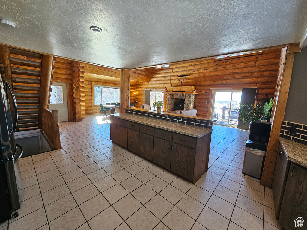 Kitchen featuring a stone fireplace, light tile patterned floors, rustic walls, and dark brown cabinetry