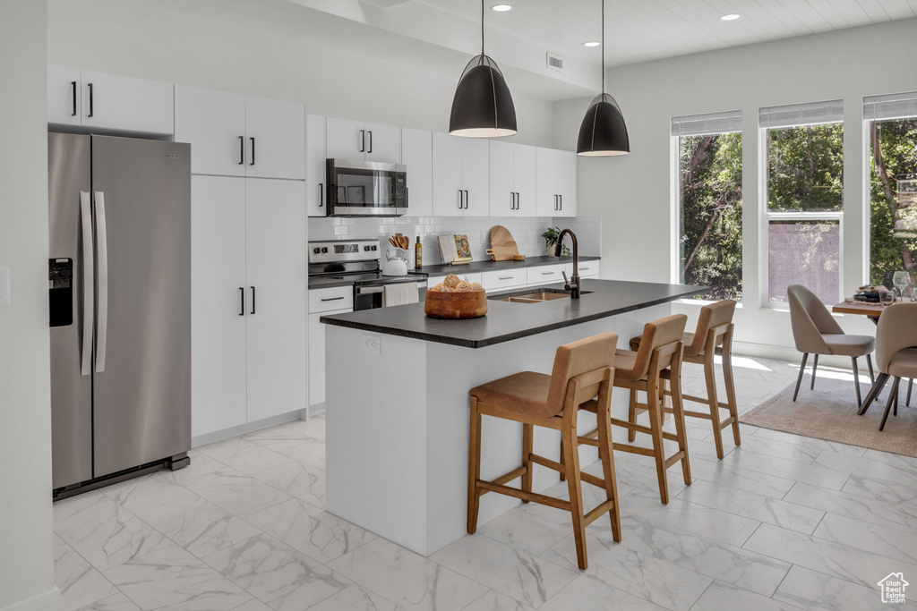 Kitchen featuring pendant lighting, sink, appliances with stainless steel finishes, white cabinetry, and an island with sink