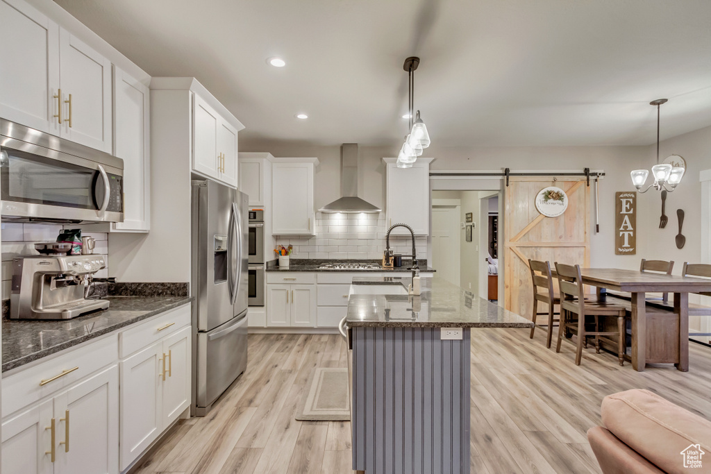 Kitchen featuring a barn door, wall chimney exhaust hood, stainless steel appliances, and hanging light fixtures