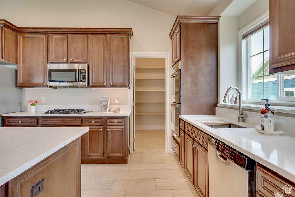 Kitchen with brown cabinets, vaulted ceiling, a sink, light stone countertops, and appliances with stainless steel finishes