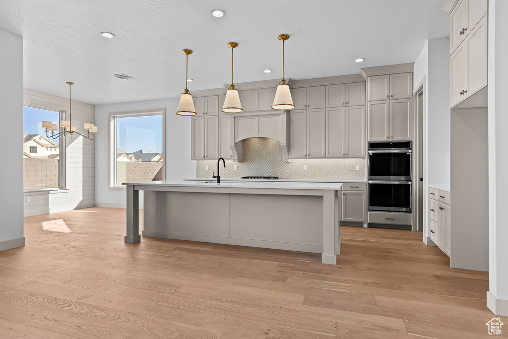 Kitchen featuring gray cabinetry, hanging light fixtures, backsplash, stainless steel double oven, and light wood-type flooring