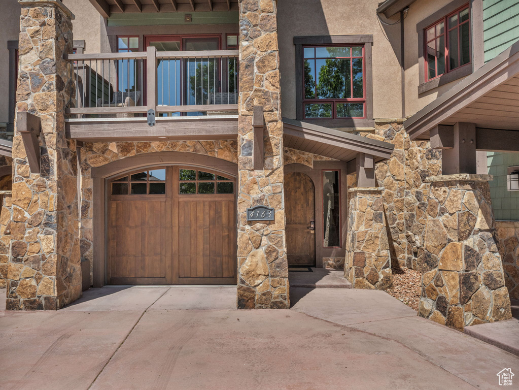 View of exterior entry with concrete driveway, stone siding, a balcony, an attached garage, and stucco siding