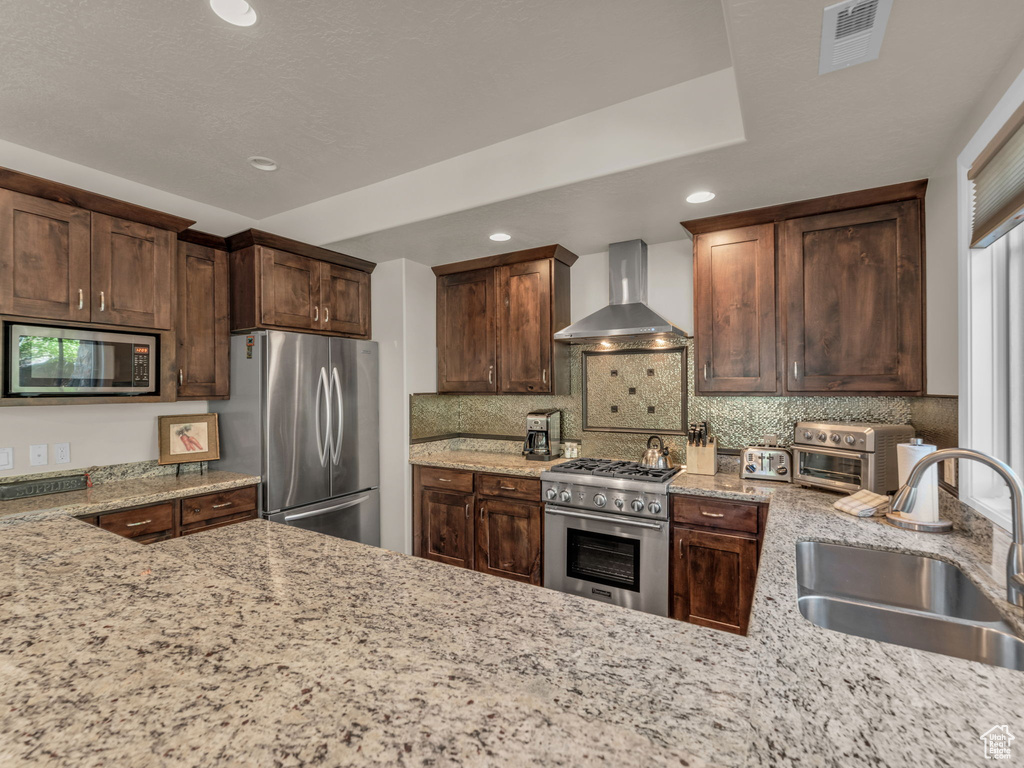 Kitchen featuring wall chimney exhaust hood, stainless steel appliances, a sink, recessed lighting, and light stone counters