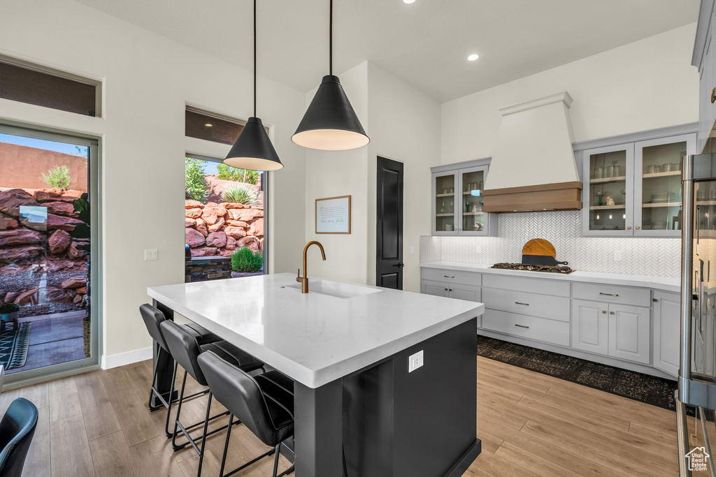 Kitchen with sink, light hardwood / wood-style floors, a center island with sink, decorative light fixtures, and custom exhaust hood