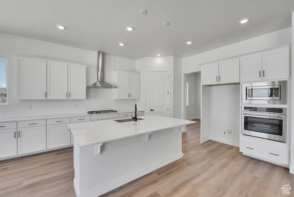 Kitchen featuring stainless steel appliances, wall chimney exhaust hood, light wood finished floors, white cabinetry, and recessed lighting