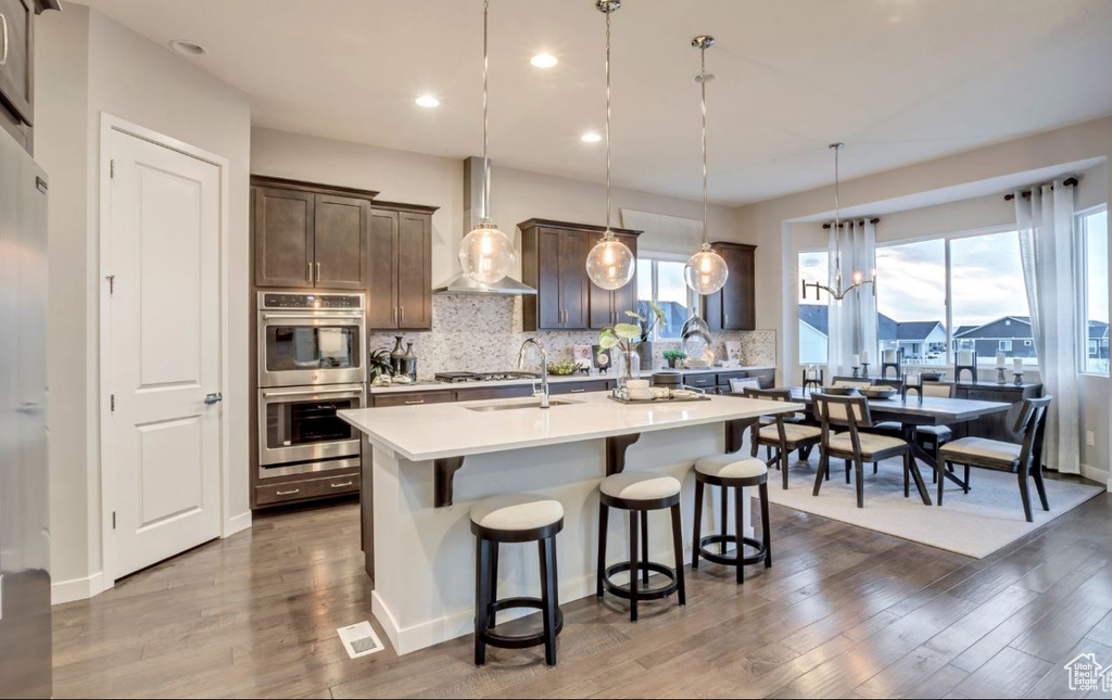 Kitchen with double oven, decorative light fixtures, an island with sink, sink, and wall chimney exhaust hood
