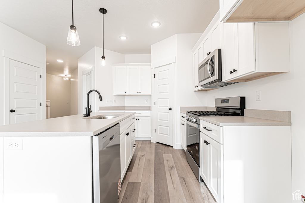 Kitchen with stainless steel appliances, light wood-style floors, a center island with sink, light countertops, and decorative light fixtures