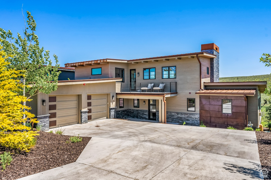 View of front of property featuring a chimney, driveway, stone siding, a balcony, and an attached garage