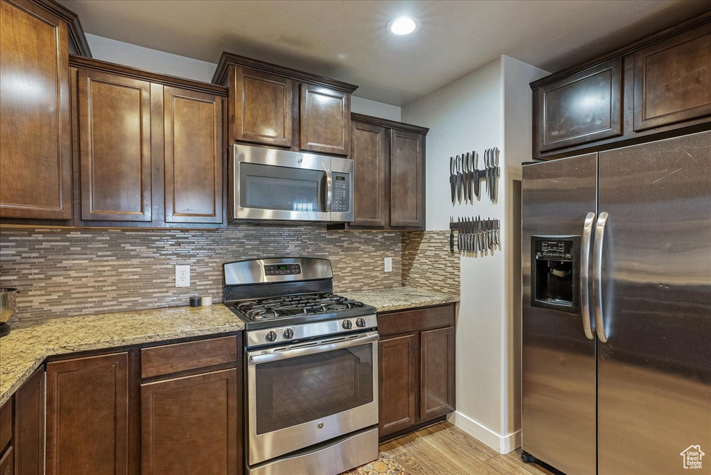 Kitchen featuring appliances with stainless steel finishes, tasteful backsplash, dark brown cabinetry, light stone counters, and light hardwood / wood-style flooring