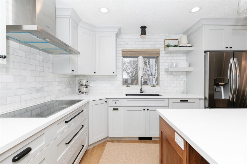 Kitchen with stainless steel refrigerator with ice dispenser, wall chimney exhaust hood, sink, black electric stovetop, and white cabinets