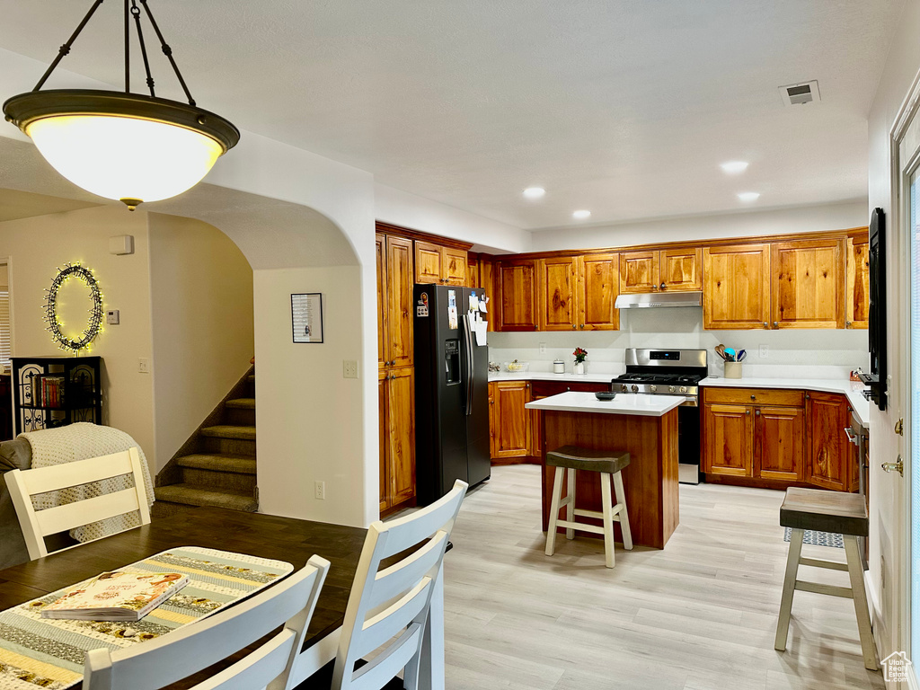 Kitchen featuring light hardwood / wood-style flooring, stainless steel gas stove, a center island, decorative light fixtures, and black refrigerator with ice dispenser