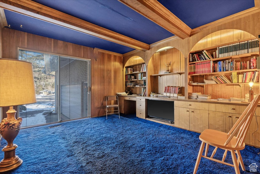 Carpeted home office featuring built in shelves, wood walls, built in desk, ornamental molding, and beamed ceiling