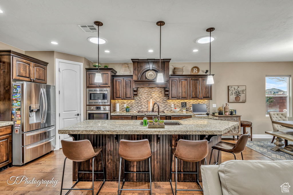 Kitchen featuring pendant lighting, a kitchen island with sink, stainless steel appliances, dark brown cabinetry, and custom range hood