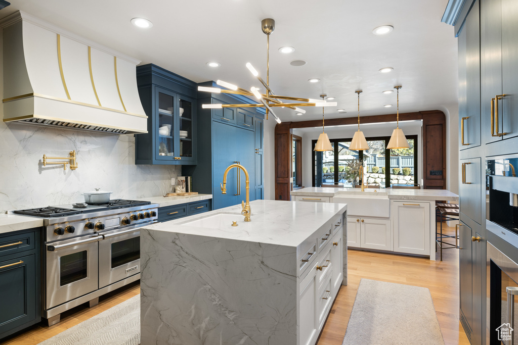 Kitchen with range with two ovens, custom exhaust hood, an island with sink, light wood finished floors, and tasteful backsplash