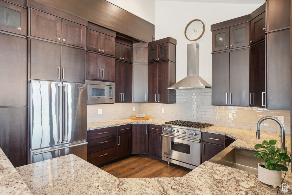Kitchen with high end appliances, wall chimney exhaust hood, a sink, dark wood-type flooring, and light stone countertops