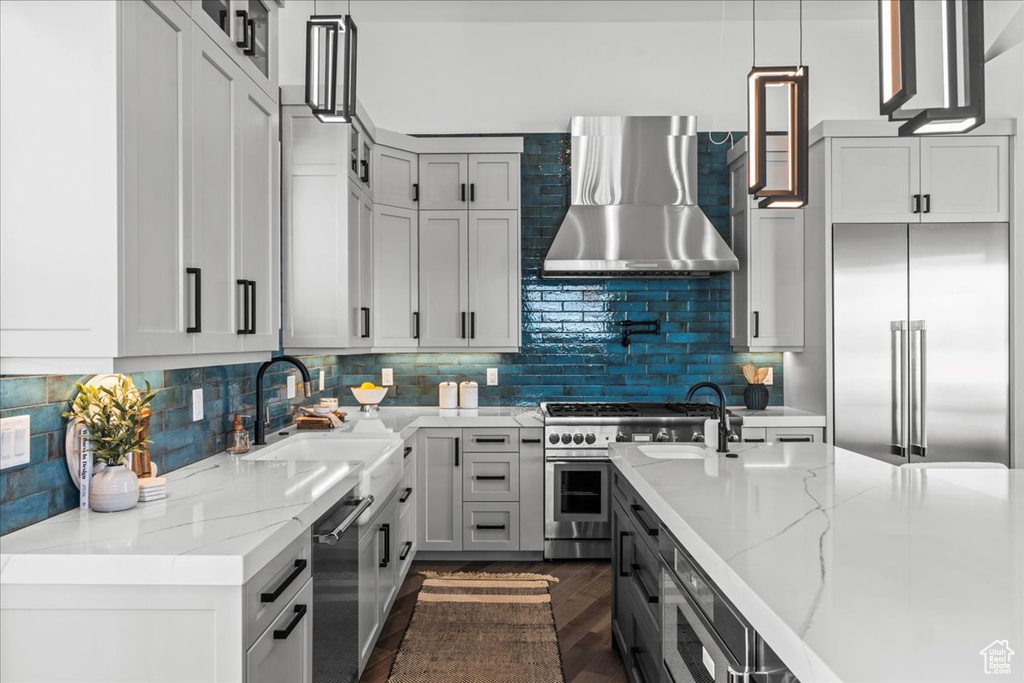 Kitchen featuring wall chimney exhaust hood, sink, light stone counters, built in appliances, and pendant lighting