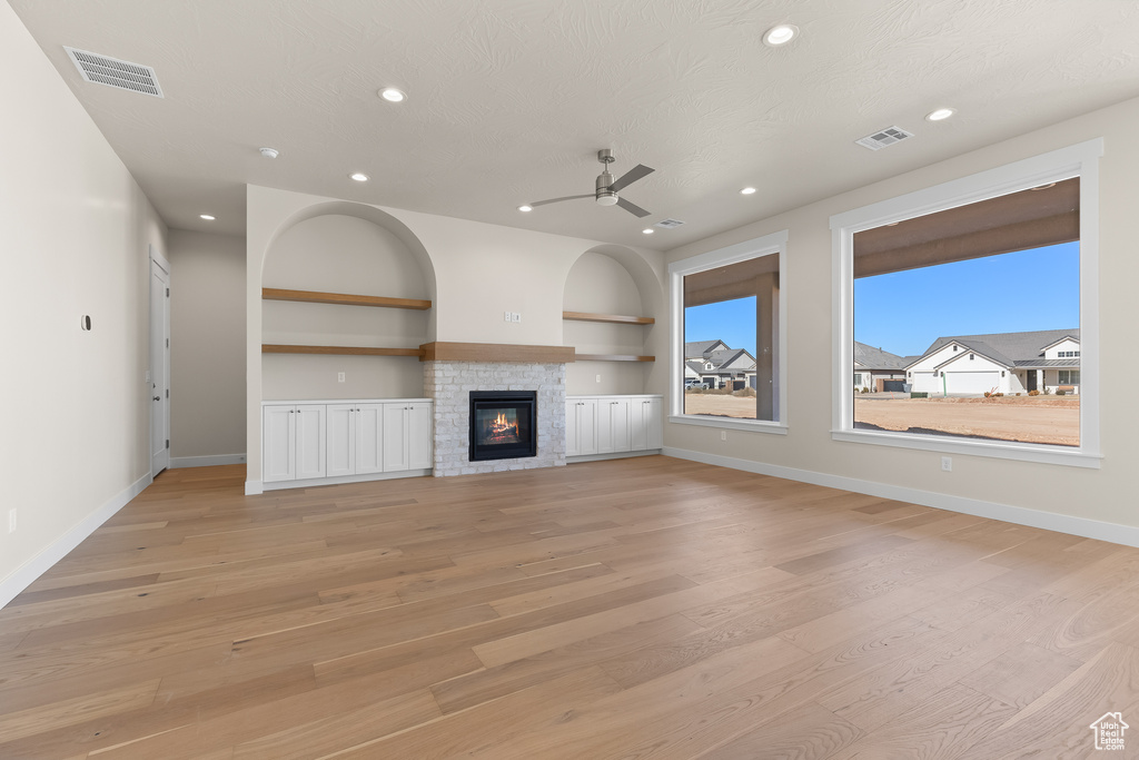 Unfurnished living room featuring a stone fireplace, ceiling fan, light hardwood / wood-style floors, a textured ceiling, and built in shelves