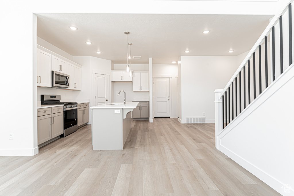 Kitchen featuring appliances with stainless steel finishes, a sink, light wood-style flooring, a center island with sink, and light countertops