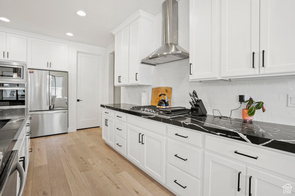 Kitchen featuring wall chimney exhaust hood, light hardwood / wood-style flooring, appliances with stainless steel finishes, dark stone counters, and white cabinets