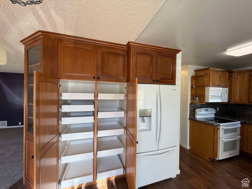 Kitchen with a textured ceiling, dark hardwood / wood-style flooring, white appliances, dark stone counters, and backsplash