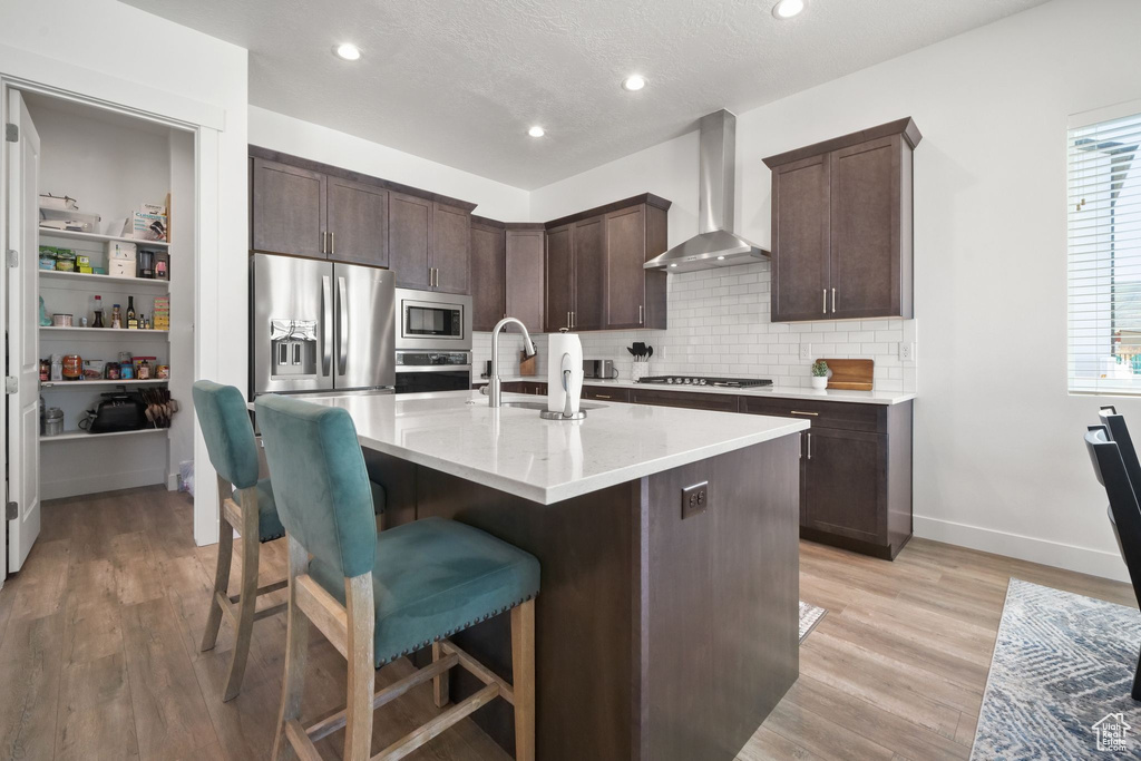 Kitchen with wall chimney range hood, sink, appliances with stainless steel finishes, a kitchen island with sink, and backsplash