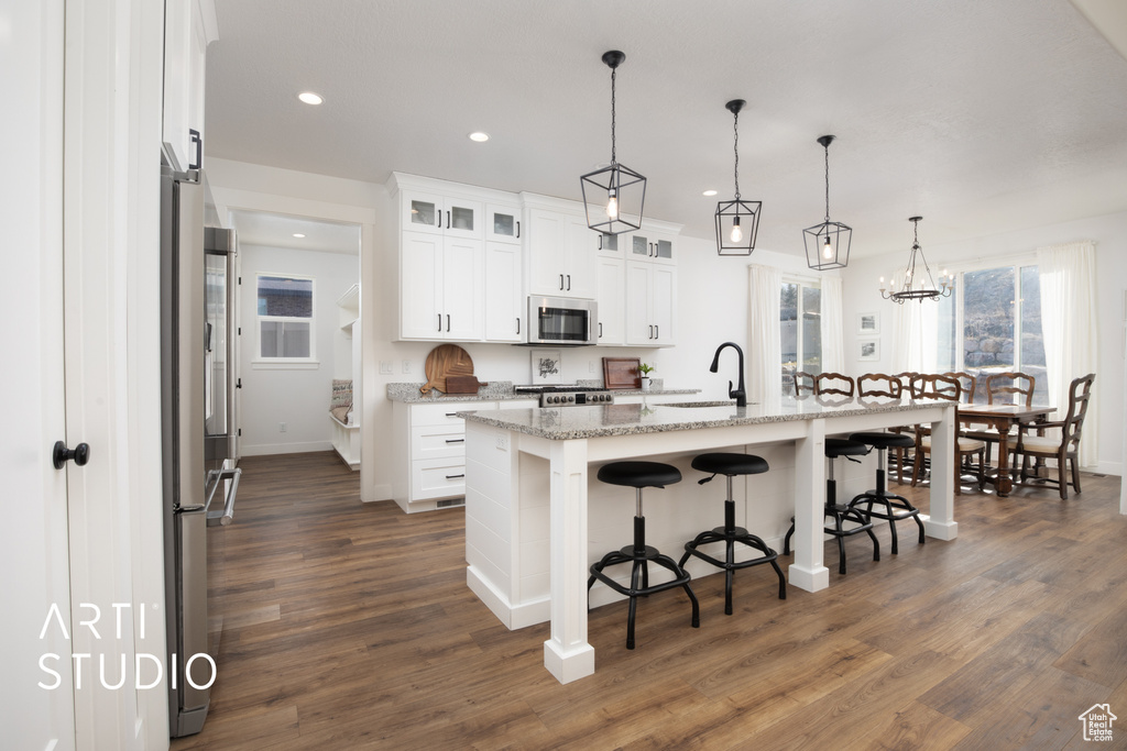 Kitchen with light stone counters, hanging light fixtures, appliances with stainless steel finishes, an island with sink, and white cabinets