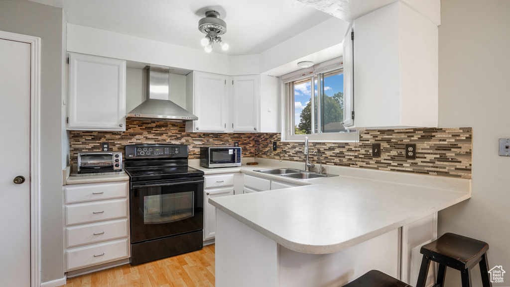 Kitchen featuring black range with electric cooktop, wall chimney range hood, a peninsula, stainless steel microwave, and a kitchen breakfast bar