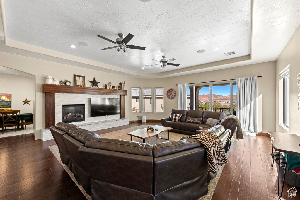 Living room with a stone fireplace, ceiling fan, a tray ceiling, dark wood-type flooring, and a textured ceiling