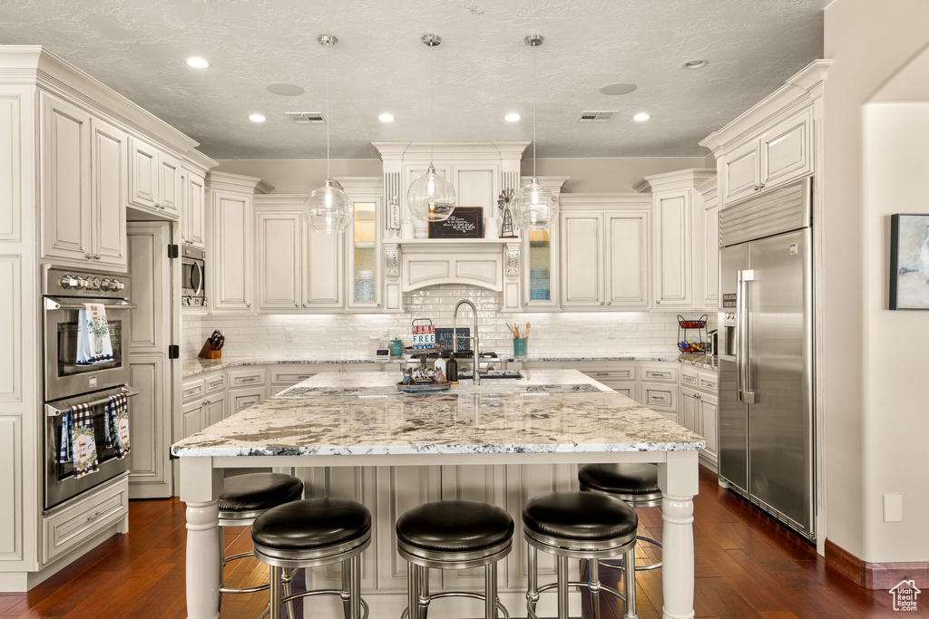 Kitchen featuring pendant lighting, a breakfast bar, appliances with stainless steel finishes, a kitchen island with sink, and light stone countertops