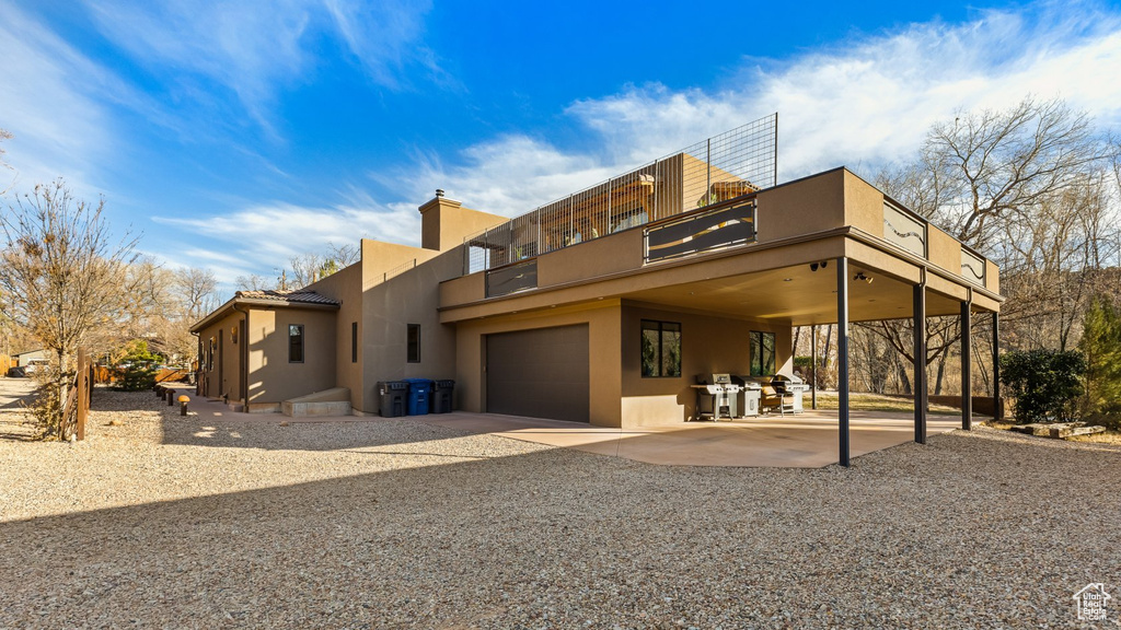 Back of property featuring stucco siding, a balcony, an attached garage, gravel driveway, and a chimney