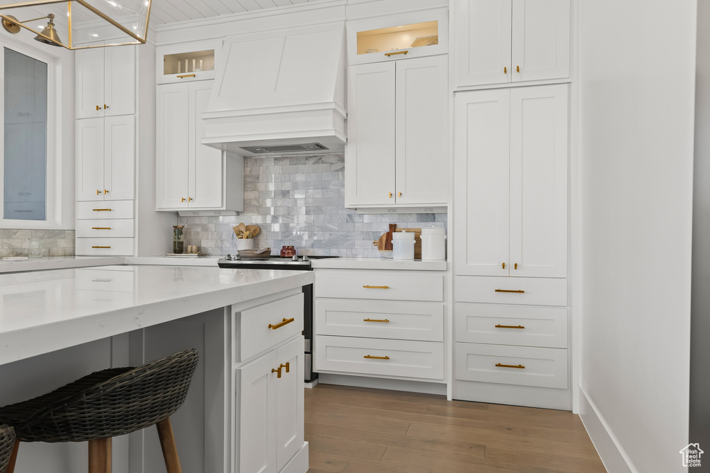 Kitchen with a breakfast bar area, premium range hood, white cabinetry, decorative backsplash, and light wood-type flooring