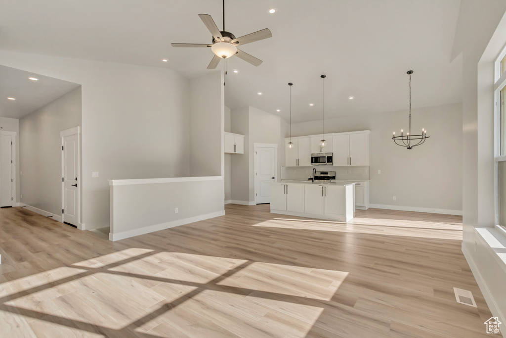 Unfurnished living room with sink, ceiling fan with notable chandelier, high vaulted ceiling, and light wood-type flooring