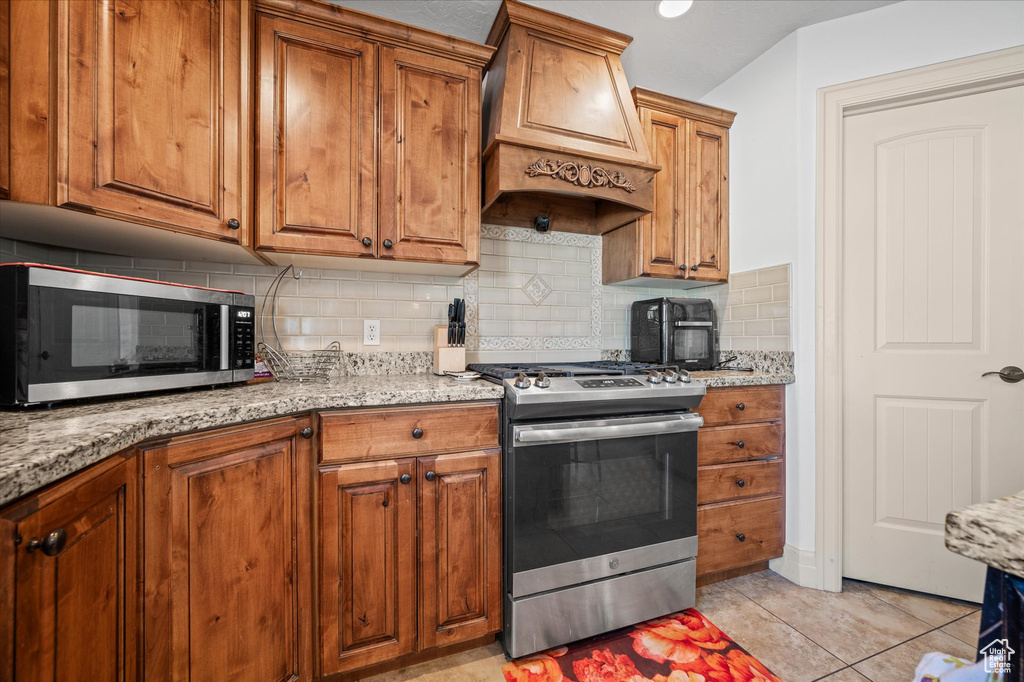 Kitchen with appliances with stainless steel finishes, decorative backsplash, custom exhaust hood, light tile patterned floors, and light stone countertops